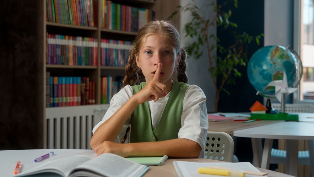 Smiling Caucasian little girl playful schoolgirl child school kid pupil learner student point finger lips mouth silence quiet noise whispering shh hush wink gesture secret indoors classroom education