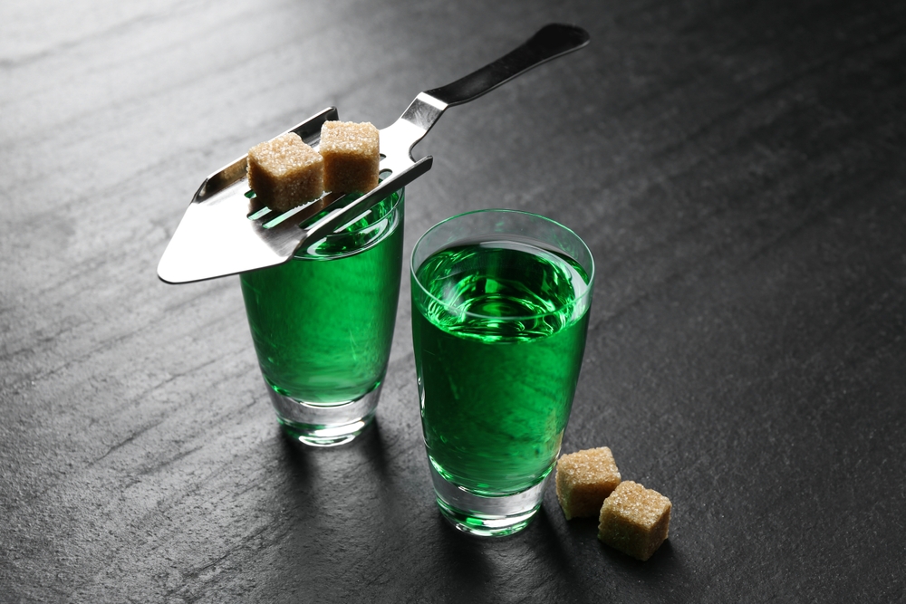 Absinthe in shot glasses, brown sugar and slotted spoon on black table, closeup
