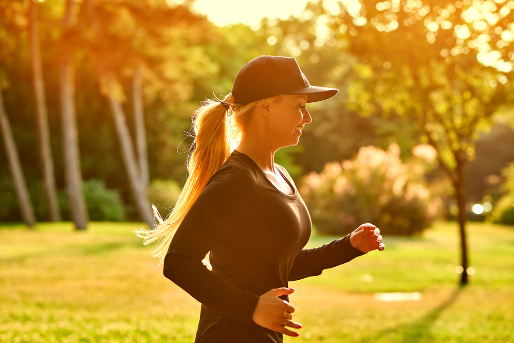 A beautiful young blonde woman wearing black clothes and baseball cap while running in a park in the sunset