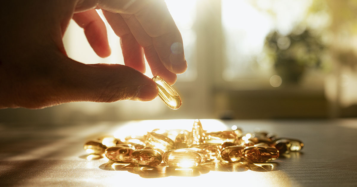 Person grabbing a soft gel capsule with sun shining in the background. Vitamin D supplement concept