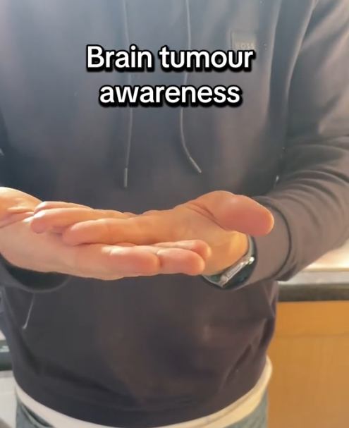 Tiktoker's boyfriend in a dark hoodie stands indoors, holding their hands out flat and gently cupped together. Bold text on the image reads "Brain tumour awareness," indicating a message of support or information about brain tumour awareness.