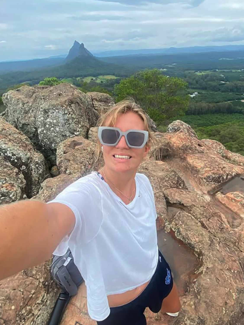 "Molly McGovern taking a selfie from a rocky overlook with a spectacular mountain vista in the background. She is wearing sunglasses, a white t-shirt, and has blonde hair. Behind her stretches a vast landscape with a distinctive mountain peak rising from lush green forests. This image captures Molly's adventurous spirit and love for travel, which she maintained throughout her battles with pheochromocytoma and paraganglioma, rare forms of cancer she fought with courage since being diagnosed at age 18.