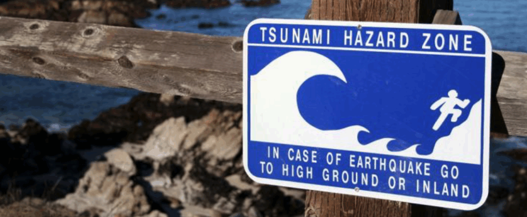 Blue hazard zone sign on wooden post telling people to go to high ground or inland when earthquakes hit near the ocean because your best warning system is your own eyes.