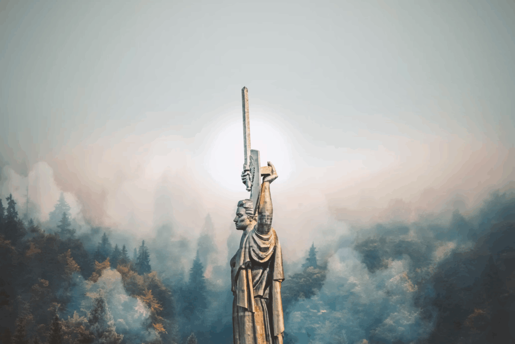 Dramatic photograph of the Motherland Monument in Ukraine emerging through misty clouds. The monumental Soviet-era statue depicts a female figure holding a sword and shield aloft, symbolizing the nation's strength and resilience. The statue is dramatically backlit with sunlight breaking through clouds, with forested areas visible below.