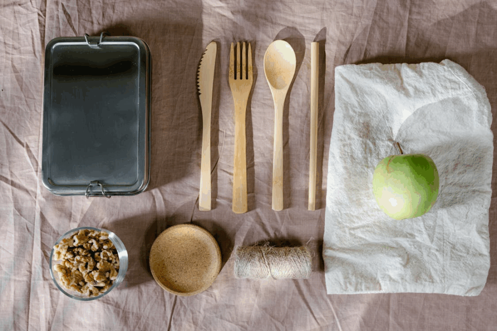 Eco-friendly food storage arrangement featuring wooden utensils, a metal container, natural fiber cloth, and a green apple, all arranged on a light fabric background as alternatives to plastic containers.