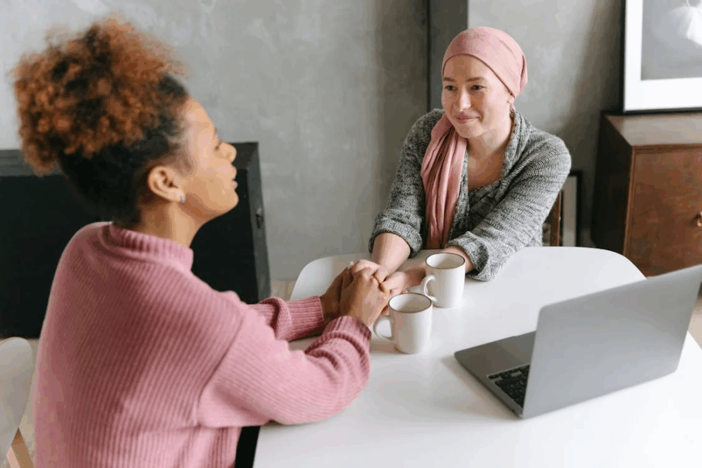 Two women sharing a meaningful conversation over coffee at a table with a laptop. One woman wearing a pink headscarf, suggesting cancer treatment, holds hands with her friend in a pink sweater, illustrating the human connection and improved quality of life that comes with less time spent in treatment.