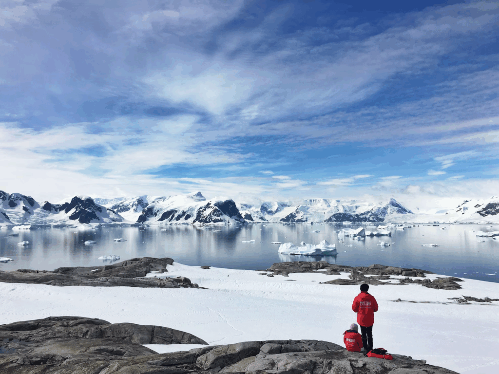 Two researchers stand on rocky terrain overlooking a vast Antarctic landscape dotted with icebergs and snow-capped mountains, illustrating the stark beauty of the continent where scientists study ice gains in Antarctica and their global implications.