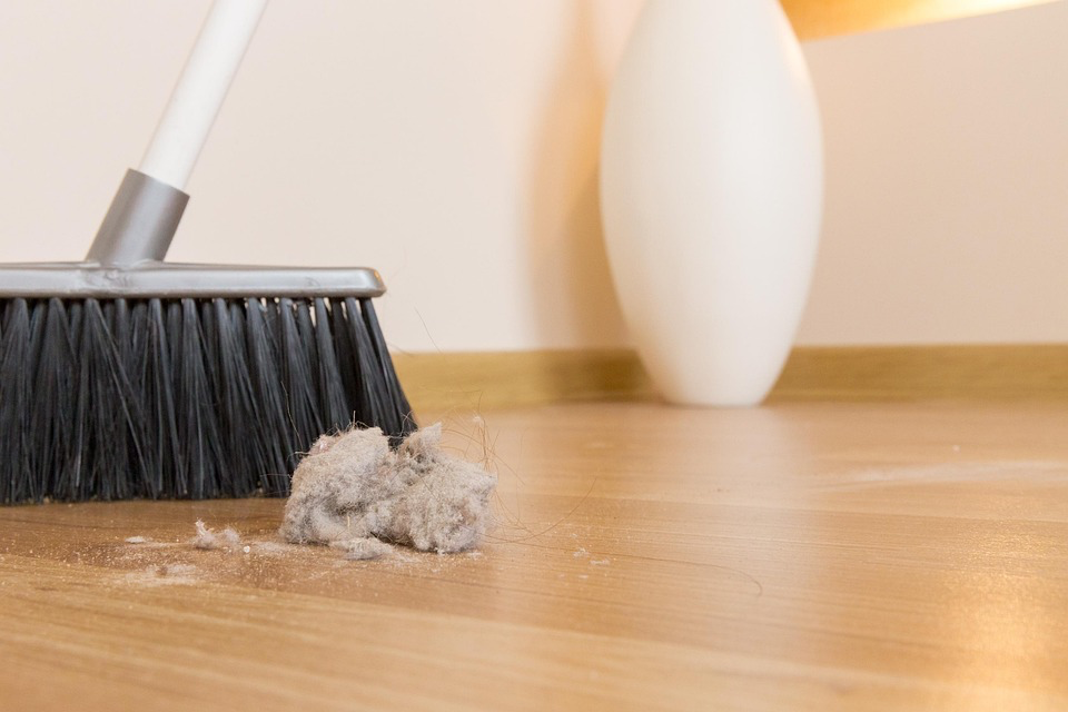 Broom sweeping a clump of dust and lint on a wooden floor, highlighting how regular cleaning helps reduce microplastic exposure by removing plastic-containing particles from home environments.