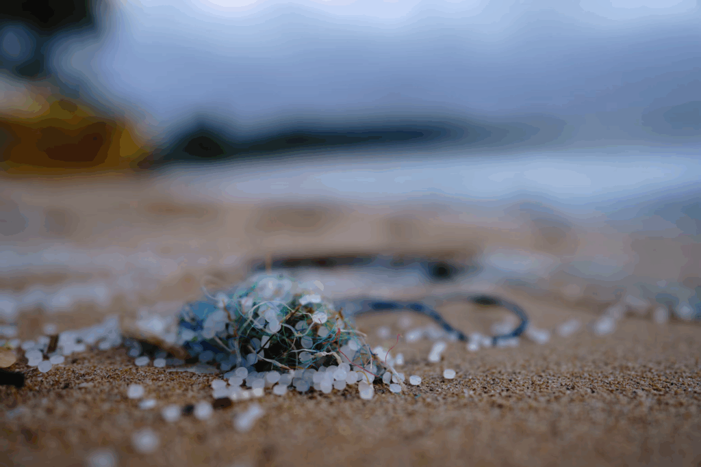 Small pile of white and blue microplastic debris mixed with sand on a beach shoreline, with blurred ocean waters in the background showing how plastic pollution affects natural environments.