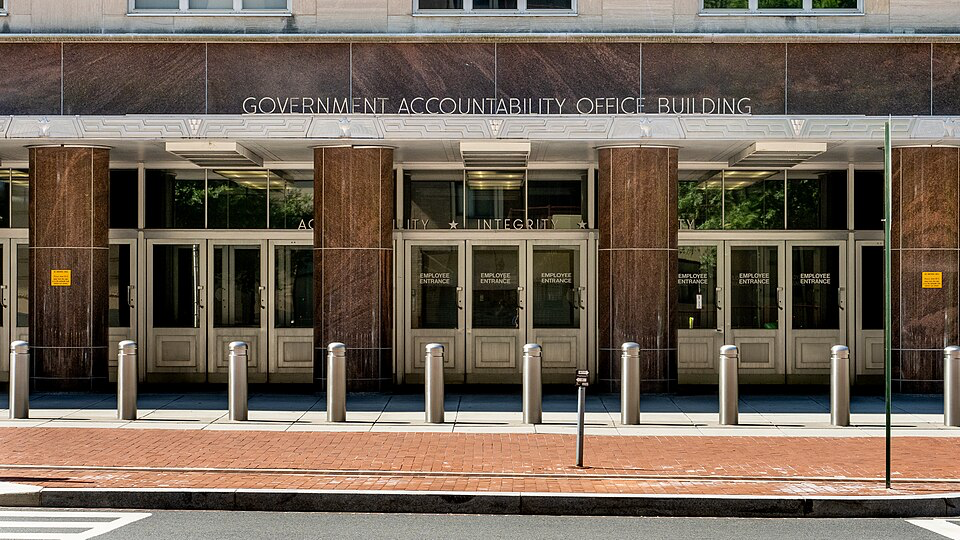 Front entrance of the Government Accountability Office building, featuring multiple glass doors with 'ACCOUNTABILITY * INTEGRITY' visible above the entrance. Security bollards line the brick sidewalk in front of this agency responsible for tracking federal spending and ensuring financial transparency.