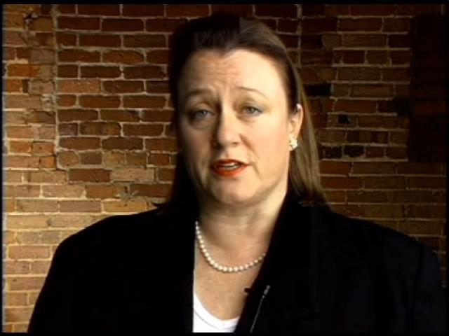 Catherine Austin Fitts, former Assistant Secretary of Housing and Urban Development during the Bush administration, speaking against a brick background while wearing a dark blazer and pearl necklace. Her claims about a secret underground city have gained attention despite lack of verification.
