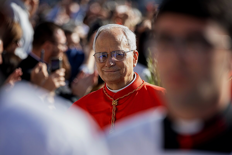 Photograph of the American Pope Leo XIV (formerly Robert Prevost) smiling at a crowd. He's wearing cardinal's red robes with a white collar and glasses. The image shows him surrounded by other clergy members in a public gathering.