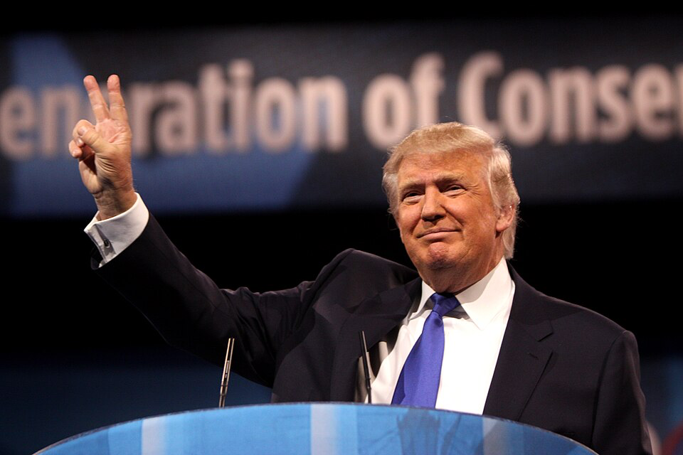 Donald Trump in a dark suit with blue tie standing at a podium, raising his hand with a peace sign gesture.