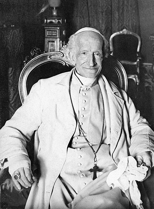 Black and white historical photograph of Pope Leo XIII seated in an ornate chair, wearing traditional white papal vestments including a zucchetto (skullcap). He has a cross pendant hanging from his neck and appears elderly with a gentle expression.