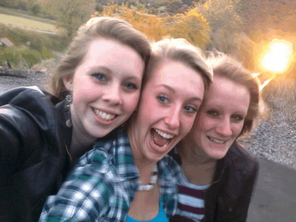 The final selfie taken by three teenage girls in Spanish Fork Canyon, Utah in 2011. Three young women with light-colored hair are smiling together near train tracks. In the upper right corner of the image, the bright headlight of an approaching second train is visible - the danger they didn't notice while focused on capturing this moment with the first train behind them. This photo was taken moments before the tragic accident described in the article.