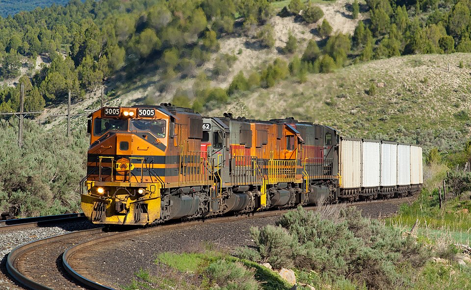 A freight train with orange and black engines numbered 5005 traveling along curved tracks through Spanish Fork, Utah. The train is moving through a rural landscape with scrubby vegetation, hills, and trees. This section of track has a light descending gradient where engines often idle, creating visible heat haze. This image illustrates the type of train and location involved in the accident described in the article.