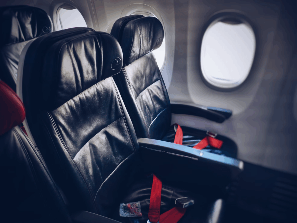 Empty airplane seats in an economy class cabin showing the standard width of seats and armrests. The leather seats are positioned next to a window, with red seat belts visible against the dark upholstery.