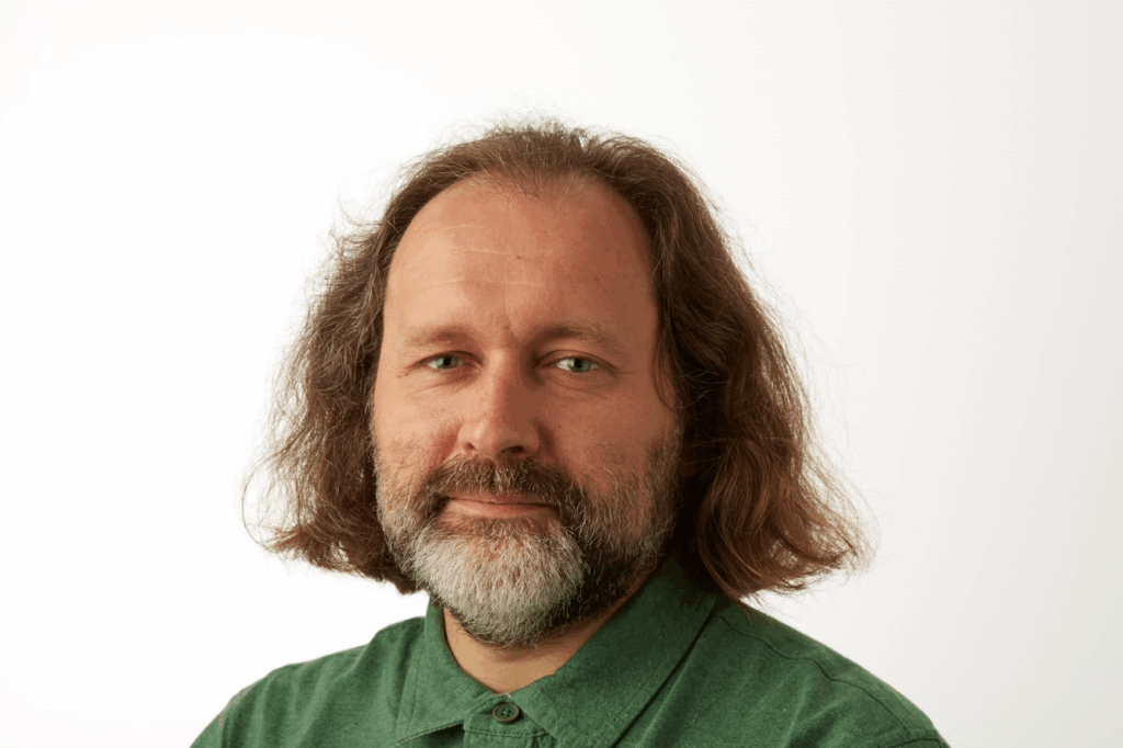 Portrait of medievalist Grigory Kessel with shoulder-length wavy brown hair and beard wearing a green collared shirt against a white background. Kessel discovered ancient biblical text using ultraviolet technology at the Austrian Academy of Sciences.
