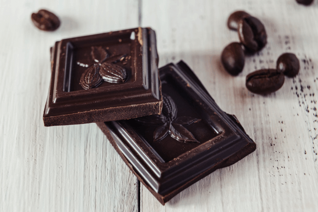 Two squares of dark chocolate with cocoa beans nearby on white wooden surface, triggers happy feelings in the brain while boosting serotonin levels.