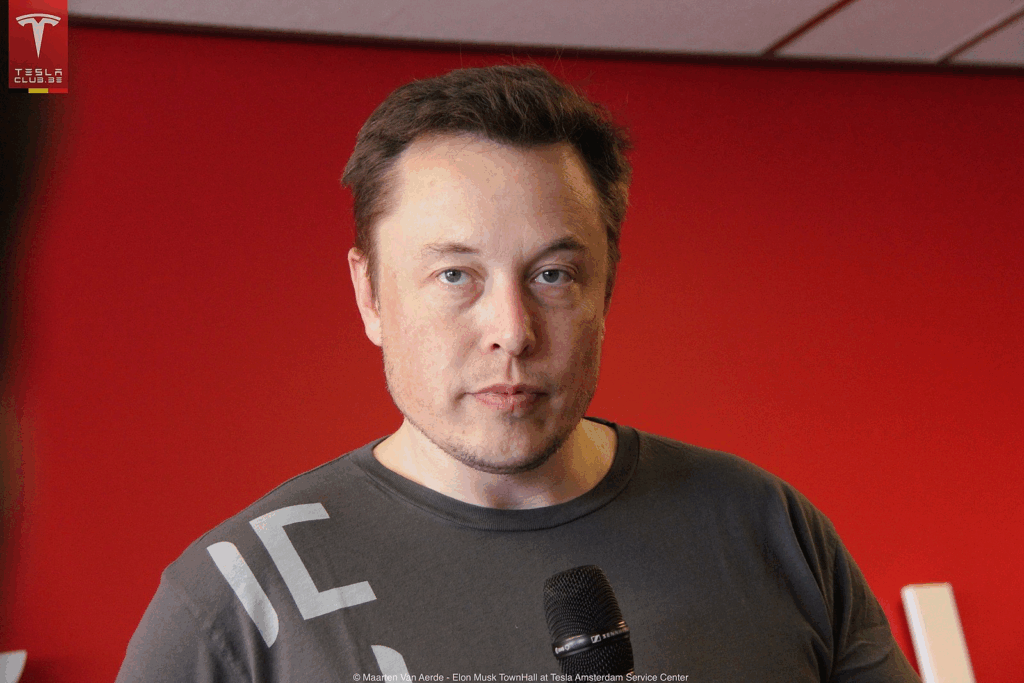 Portrait of Elon Musk with short brown hair wearing a dark gray t-shirt against a red background. The CEO and founder of SpaceX and Tesla is shown at what appears to be a Tesla event, as indicated by the Tesla Club logo in the corner.