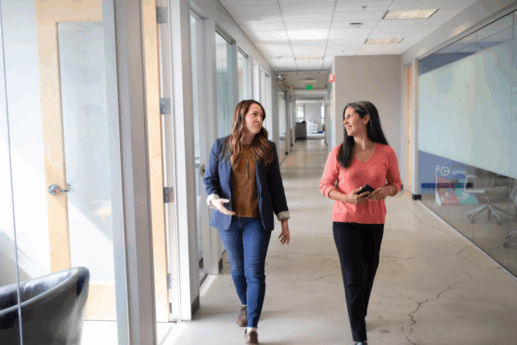 Two women walking and talking in a office hallway, demonstrating sneaky ways to walk more by taking walking meetings and moving around your workplace instead of sitting all day.