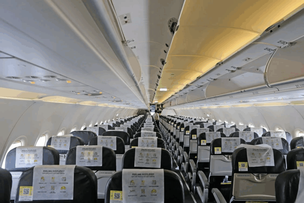 Black and white photograph of an airplane cabin interior taken from a passenger's perspective looking forward down the aisle. Multiple rows of seats are filled with passengers, demonstrating the tight seating arrangements that can be challenging for plus-sized passengers, with the curved ceiling structure of the aircraft clearly visible.