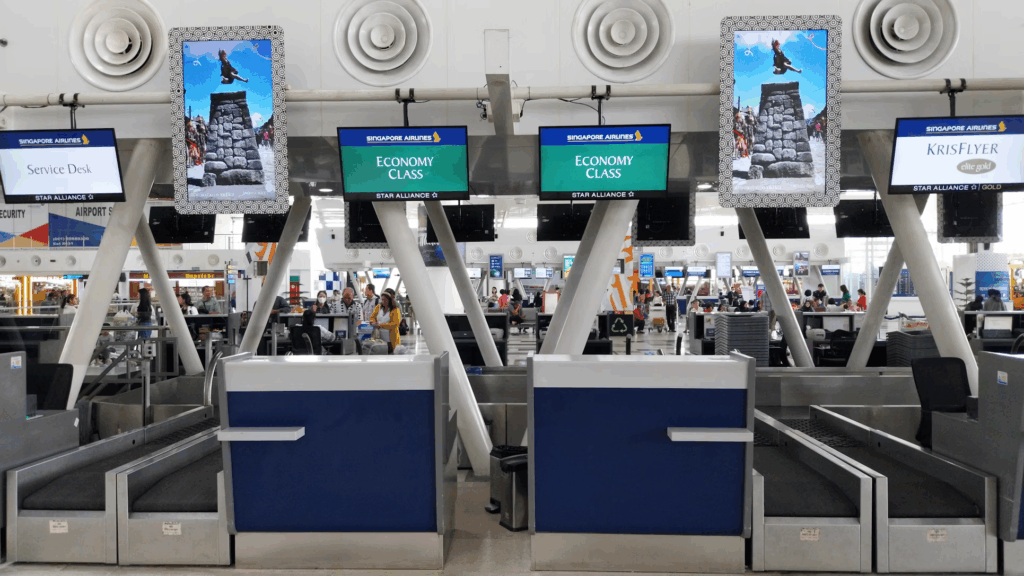 Airport check-in area showing multiple self-service kiosks with blue and white Singapore Airlines branding. Digital screens display "ECONOMY CLASS" and decorative panels above feature images of landmarks. The modern terminal setting includes white structural elements and passengers can be seen in the background using the check-in facilities.
