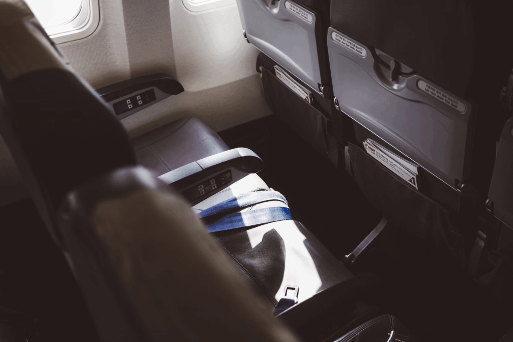 A close-up view of an airplane passenger seat with a white and blue striped safety information card or magazine placed on the dark upholstery. The seat appears to be in a premium cabin with modern gray interior paneling and windows visible in the background, highlighting the space constraints that affect plus-sized passengers.