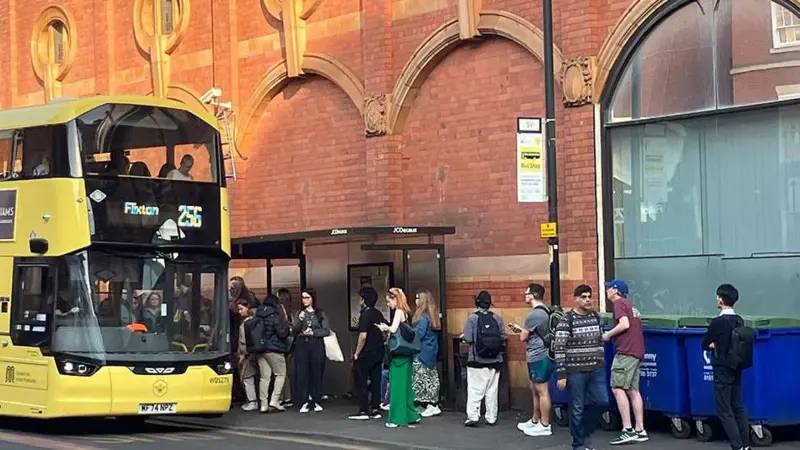 Passengers queue for a yellow double-decker bus in Manchester following the power outage that halted tram services. People with backpacks and bags stand in line outside a red brick building with arched features, able to use their tram tickets on local buses while the transport system recovers.