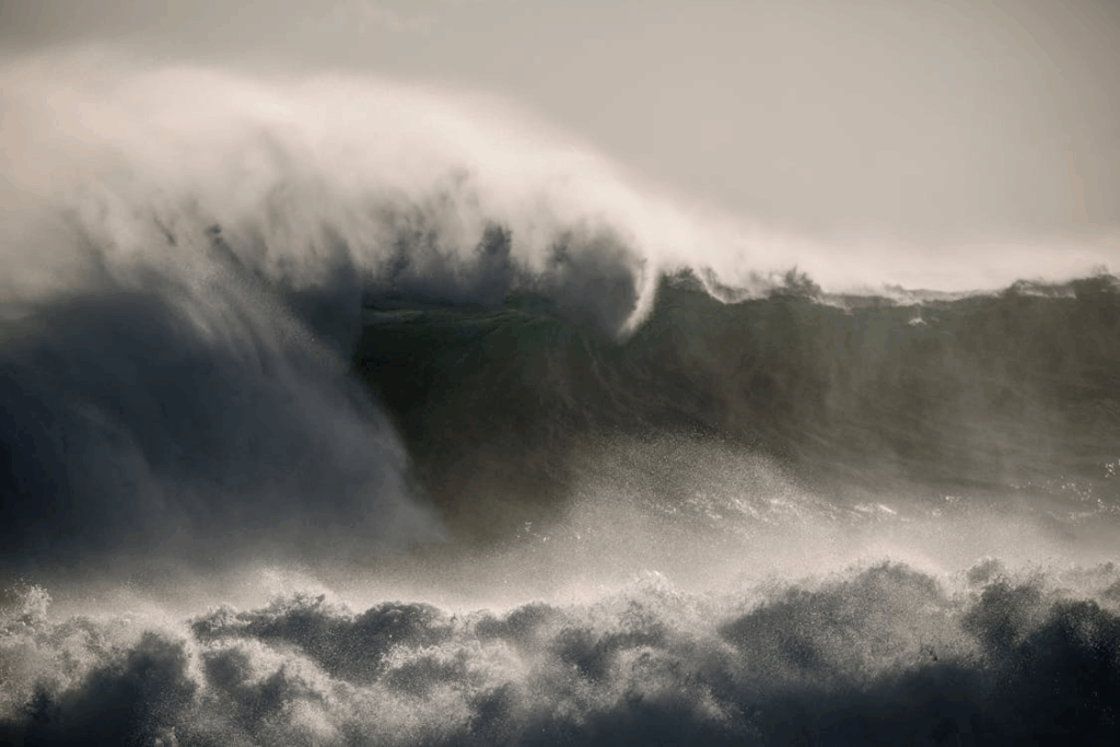 Massive ocean waves crash with tremendous force, their white foam and spray captured mid-motion against a dark, stormy sky. The turbulent seawater demonstrates the kind of devastating tsunami power that Ryo Tatsuki claims will strike the Pacific region in her controversial July 2025 prediction.
