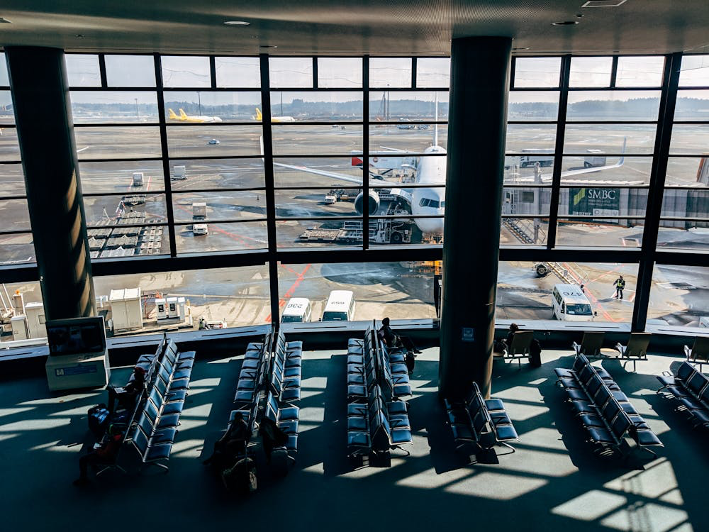 Empty airport terminal seating faces large windows overlooking busy tarmac operations, with aircraft and ground vehicles visible outside. The vacant chairs reflect the sharp decline in travel bookings to Japan as tourists cancel trips due to widespread fear over the predicted disaster.