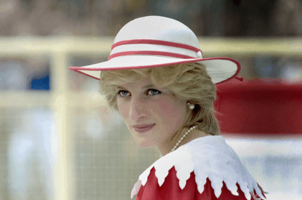 Princess Diana wearing an elegant white hat with red trim and matching white outfit with red geometric details looks directly at the camera with her characteristic gentle smile. Her blonde hair frames her face as pearl earrings catch the light, capturing the beloved royal's timeless grace and beauty that captivated the world before her tragic death in 1997.