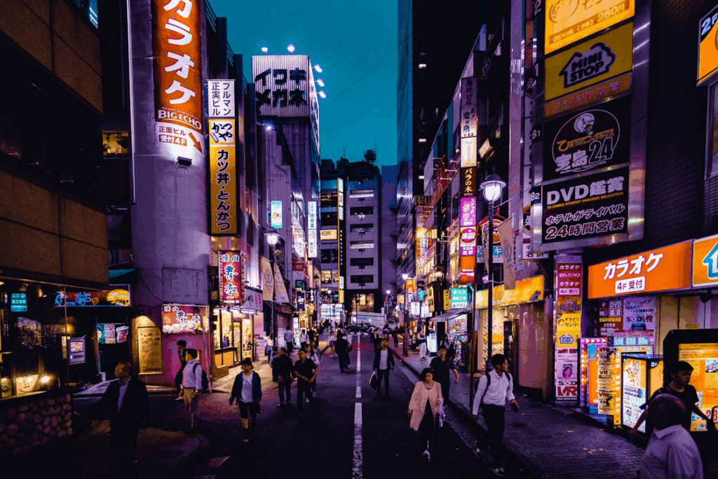 A busy Japanese street scene at dusk shows pedestrians walking between tall buildings adorned with bright neon signs and colorful advertisements. The lively urban atmosphere captures the vibrant, safe reality of modern Japan that contradicts the fear-mongering surrounding unscientific disaster predictions.