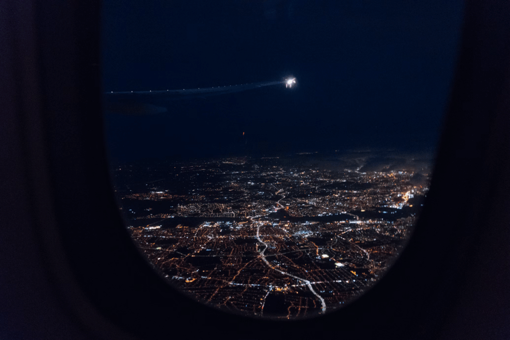 View from an airplane window showing a city illuminated at night, with streetlights creating patterns across the urban landscape. This aerial perspective of London demonstrates how electrical grids power modern cities, highlighting the vulnerability of urban centers to power outage events.