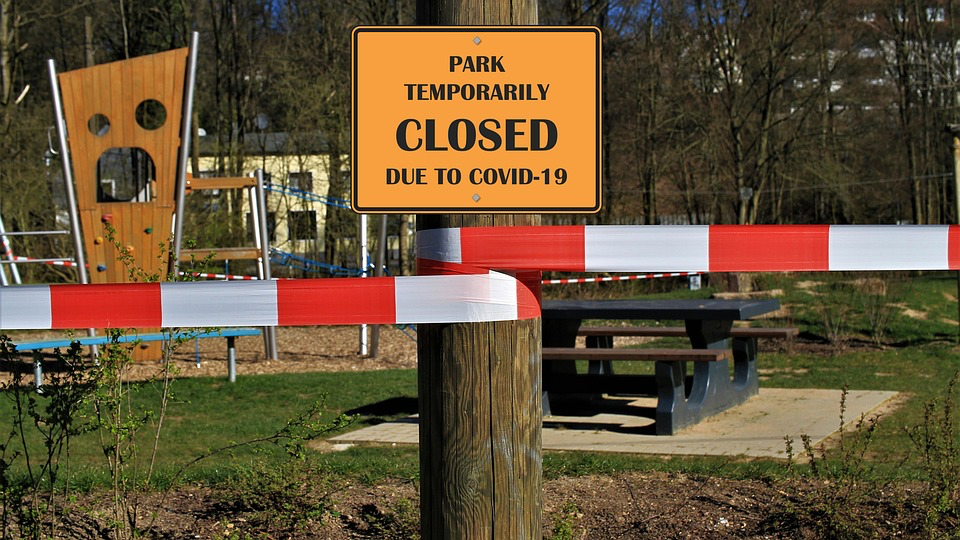 Orange sign reading "PARK TEMPORARILY CLOSED DUE TO COVID-19" with barrier tape, showing how lockdowns prevented disease spread but delayed the expected whooping cough outbreak.