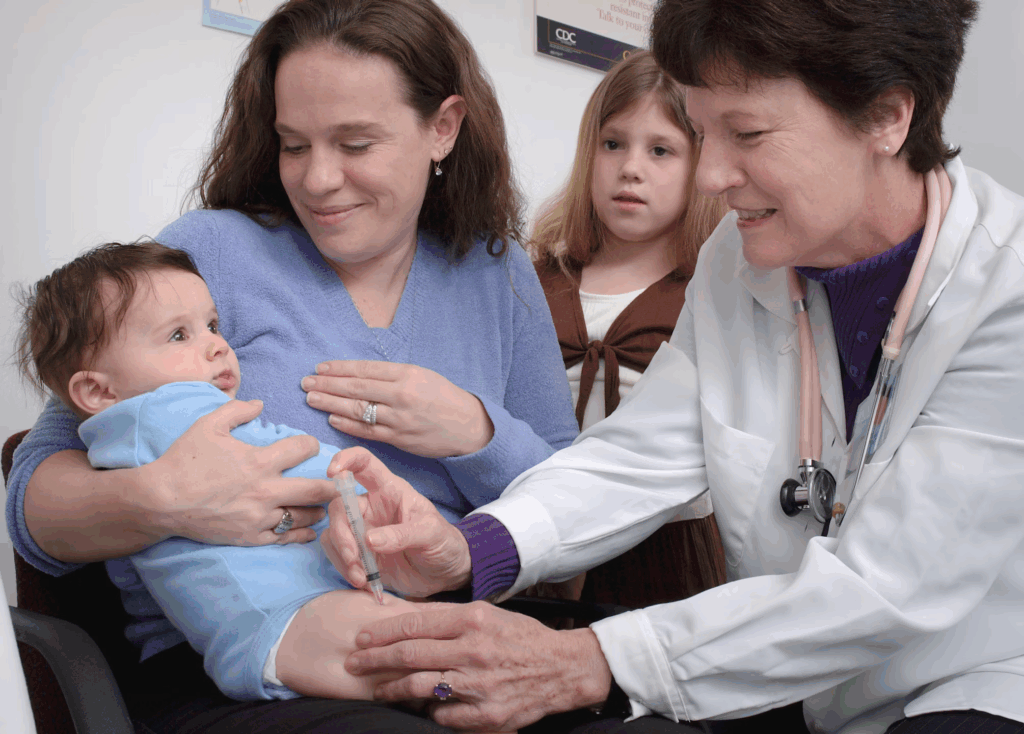 Doctor administers injection to infant held by mother while family observes in medical office during vaccination appointment.
