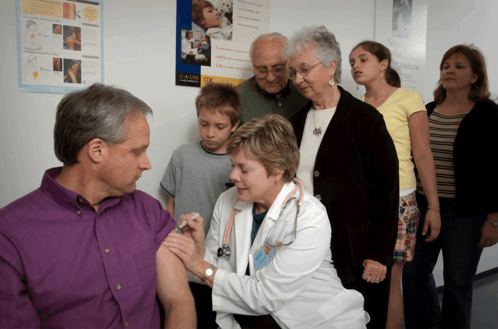 Doctor administers vaccination to adult patient while family members observe in medical clinic setting.