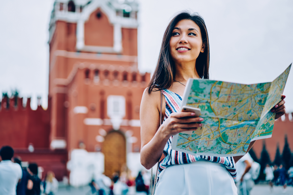 Attractive asian female traveler standing on red square in Moscow enjoying sightseeing tour with map,cheerful woman tourist getting to location near Kremlin on background planning next route in Moscow