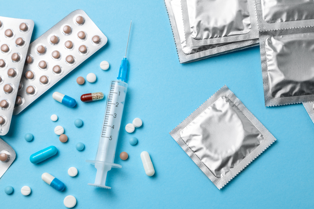 Birth control pills, an injection syringe and condom in a package on blue background. The concept of choosing method of contraception, birth control pills or condom.
