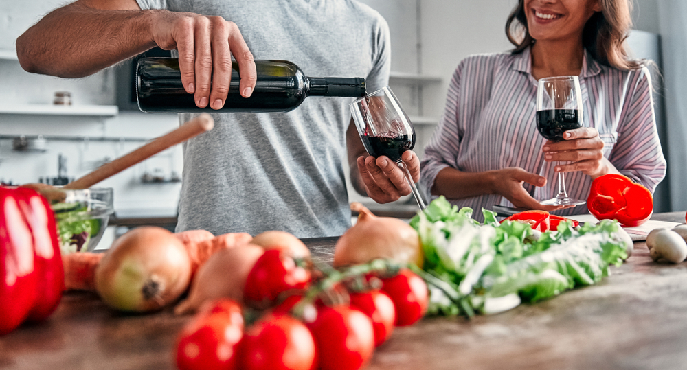 Romantic couple is cooking on kitchen. Handsome man pours wine into glasses and attractive young woman are having fun together while making salad. Healthy lifestyle concept.