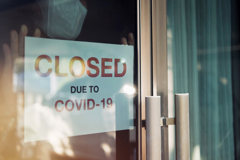 Business office or store shop is closed, bankrupt business due to the effect of novel Coronavirus (COVID-19) pandemic. Unidentified person wearing mask hanging closed sign in background on front door.
