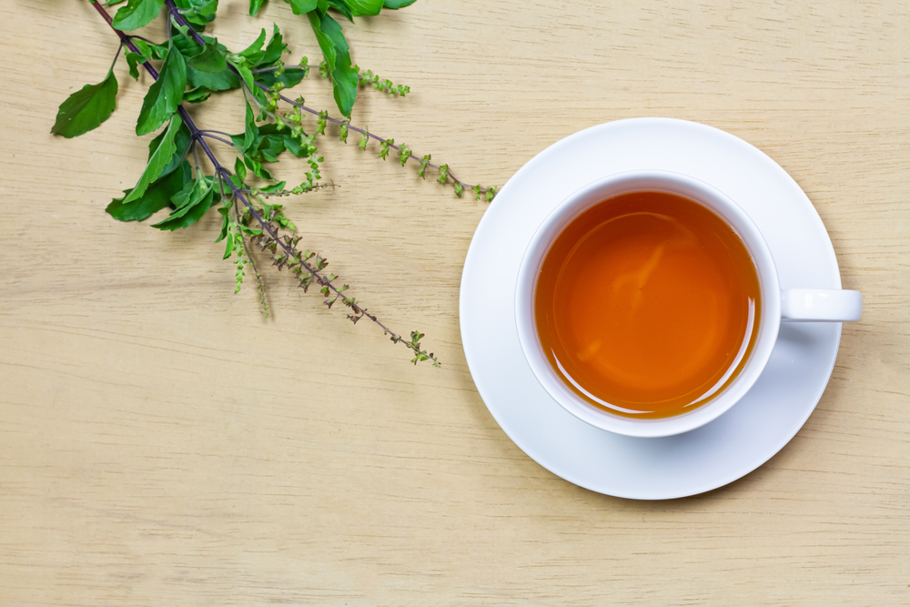 Tulsi or holy basil tea in white cup with fresh tulsi branch on wooden background top view. Ayurvedic medicine in India.