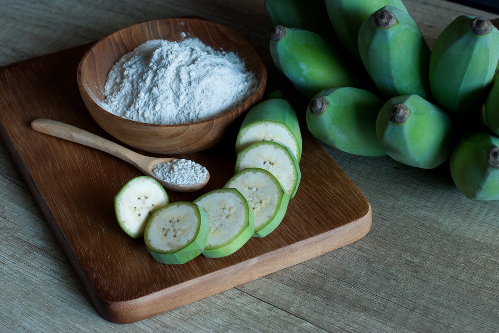 Banana flour with slice and whole green raw bananas on a wooden cutting board, prebiotic food for gut health.