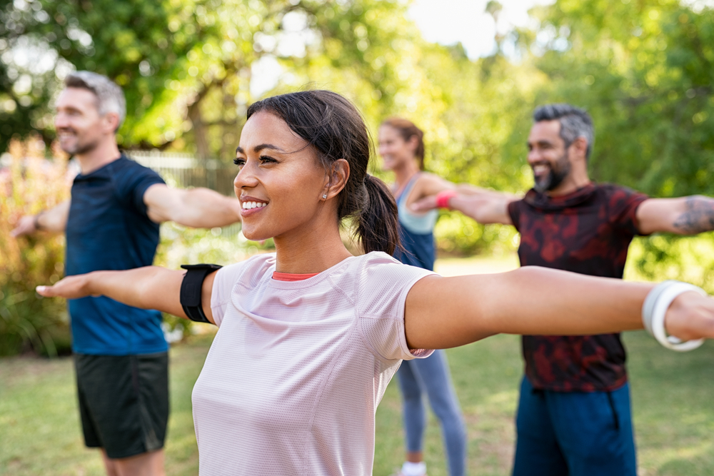 Group of multiethnic mature people stretching arms outdoor. Middle aged yoga class doing breathing exercise at park. Beautifil women and fit men doing breath exercise together with outstretched arms. 
