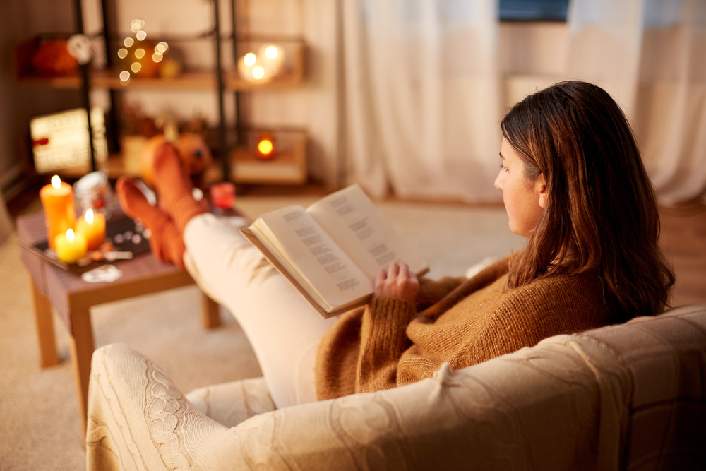 halloween, holidays and leisure concept - young woman reading book and resting her feet on table at cozy home