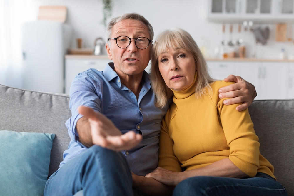 Discontented Senior Couple Talking To Camera Disapproving Criticizing Something Sitting Together On Sofa At Home. Displeased Customers. Communication Problems With Older Parents