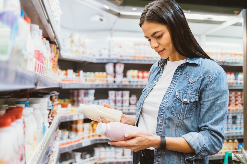 Portrait young woman in supermarket holding two bottles of yogurt reads labels compares chooses healthy dairy food