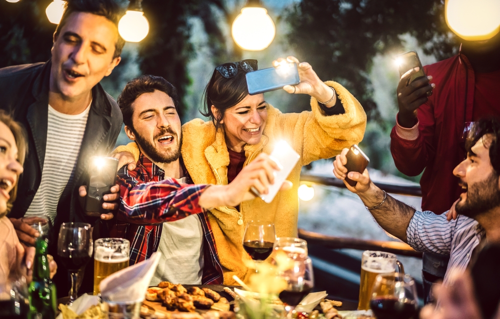 Mixed age men and women having fun drinking out at wine diner - Food beverage life style concept on happy trendy people enjoying time together at villa patio - Warm filter with bulb string lights halo