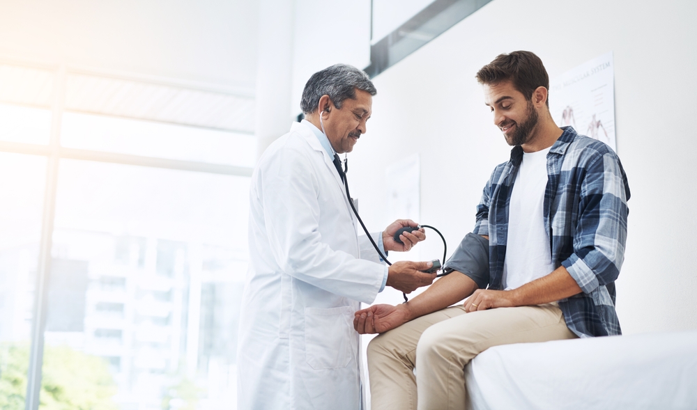 Healthcare, patient and doctor with blood pressure test, help and wellness checkup of person at hospital. Medicine, health care expert and Indian man in doctors office consulting medical professional
