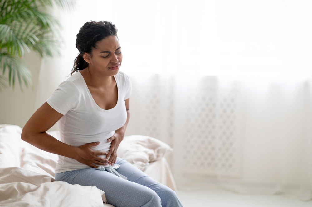 Young black woman suffering stomach pain in the morning, sitting on bed in bedroom and touching belly, african american lady in pajamas feeling unwell at home, having menstruation cramps, copy space